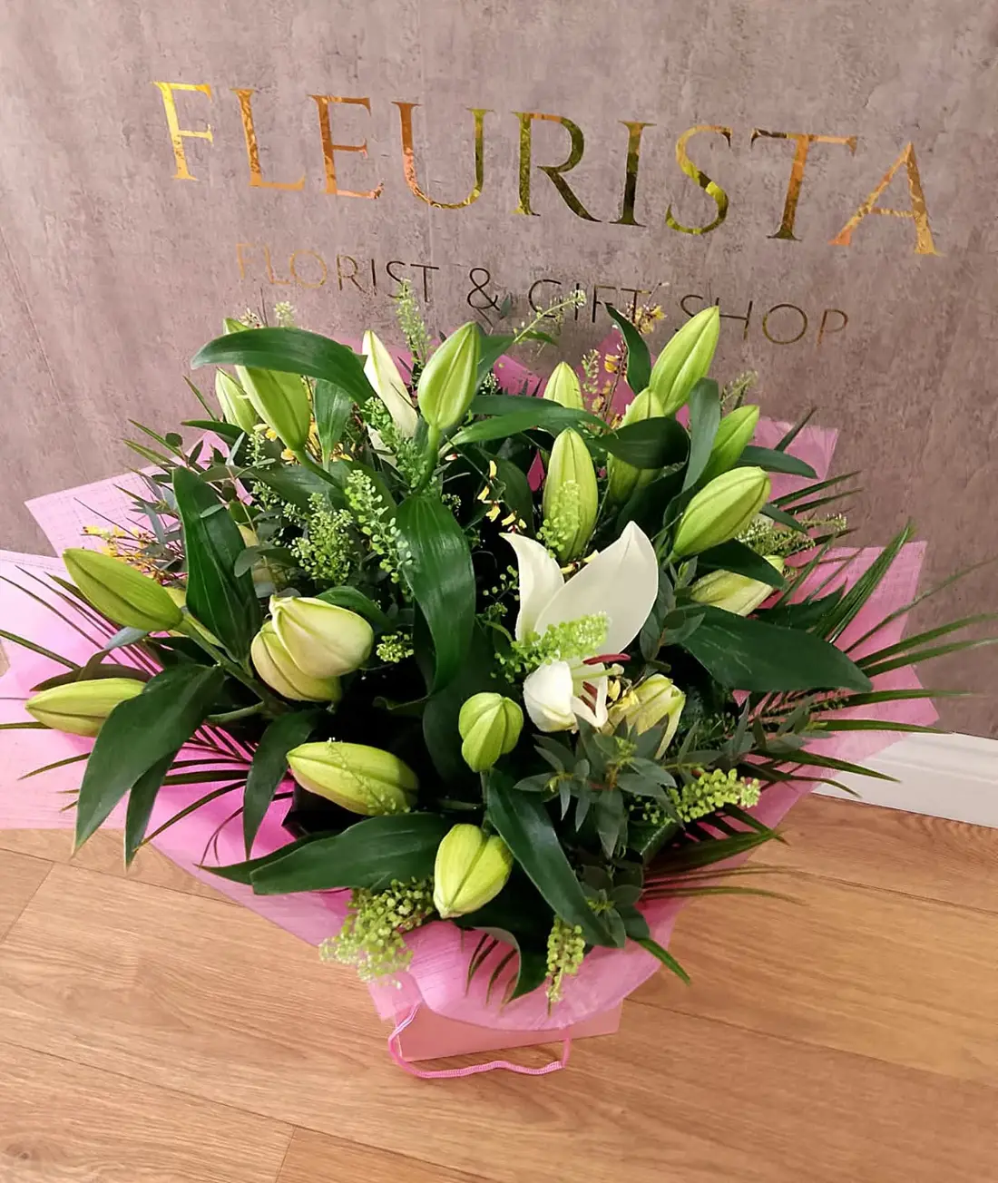 White lily bouquet wrapped in pink paper against a grey wall bearing gold foil FLEURISTA FLORIST AND GIFT SHOP signage