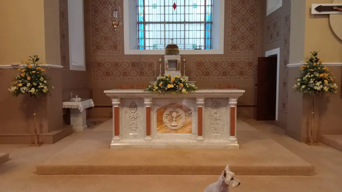 White and yellow lily altar arrangements either side of a marble church altar beneath stained glass windows for a wedding service