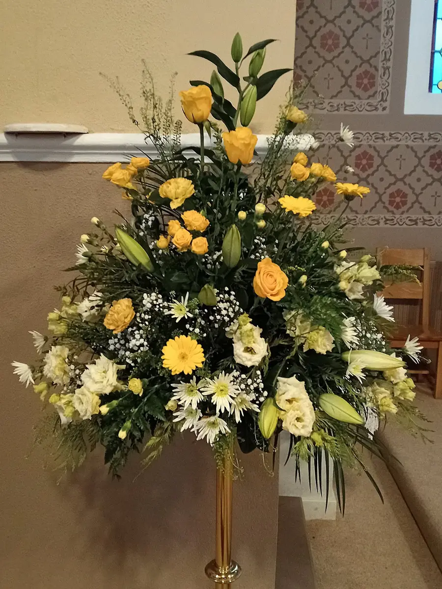 Tall brass pedestal holding a wedding arrangement of yellow roses, lilies, chrysanthemums and white flowers beside a stained glass window